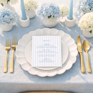 May include: A beautifully set table with white plates, gold cutlery, and a menu card. The table is decorated with blue and white hydrangeas in white vases, and light blue candles. The menu card has a blue and white striped design.
