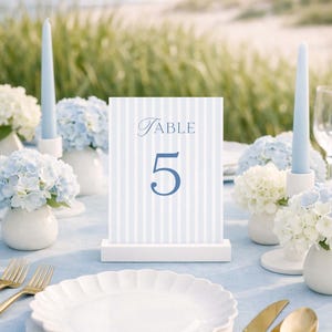 May include: A beautifully set table with a light blue tablecloth, white plates, and gold cutlery. A table number card with the word "Table" and the number "5" is displayed. Blue and white flowers and candles complete the elegant setting.