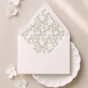 Könnte beinhalten: Ein weißer Umschlag mit geöffneter Klappe, der ein gemustertes Innenfutter mit grünen Ranken, braunen Zweigen und kleinen weißen Blüten zeigt. Der Umschlag liegt auf einem weißen Teller mit gewelltem Rand, begleitet von vier weißen Hortensienblüten.