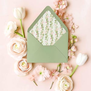 May include: A sage green envelope with an open flap revealing a decorative interior liner featuring a vertical pattern of pink roses, green leaves, and thin stripes, surrounded by an arrangement of white and pale pink ranunculus and tulip flowers.