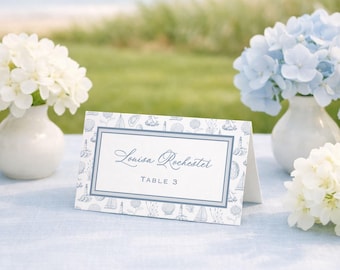Può includere: Un segnaposto rettangolare piegato con un centro bianco che riporta il testo Louisa Rochester e TABLE 3 in inchiostro blu, circondato da un bordo blu e da un motivo di illustrazioni nautiche blu tra cui barche a vela, conchiglie e fari.