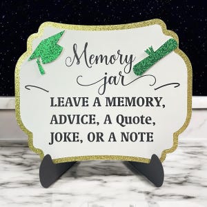 May include: A white sign with gold glitter trim and green glitter accents. The sign reads "Memory jar" and "Leave a memory, advice, a quote, joke, or a note".