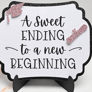 May include: A white sign with black text and a black border reads "A Sweet Ending to a New Beginning". The sign has a pink glitter graduation cap and diploma on it. The sign is on a black stand.