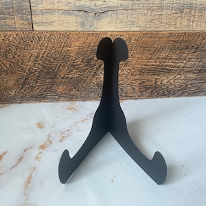 May include: A black metal display stand with a three-legged design. The stand has a curved top and is designed to hold a plate or other decorative item. The stand is shown on a white surface with a wood-paneled background.