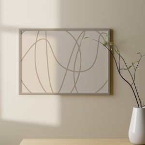 May include: A framed abstract print with brown lines on a white background hangs on a beige wall above a wooden table. A white vase with a small branch with green leaves sits on the table.
