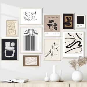 May include: A gallery wall with nine framed prints featuring abstract line art, black and white illustrations, and botanical designs. The prints are arranged in a symmetrical pattern on a white wall.