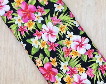 May include: A bolt of fabric featuring a black background with a vibrant tropical floral pattern. The design includes large pink and white hibiscus flowers, smaller yellow and white plumeria blossoms, and bright green palm leaves throughout.