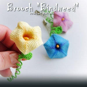 May include: Crochet pattern for a bindweed flower brooch. The brooch is made with yellow, blue, and pink yarn. The brooch is attached to a green stem.