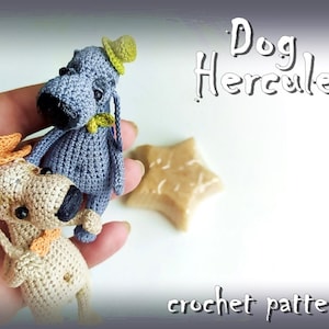 May include: Two crocheted dog toys, one grey with a green hat and one beige with an orange hat. The toys are on a hand and the text "Dog Hercule crochet pattern" is below them.