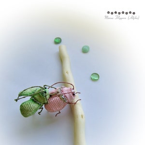 May include: Two crocheted bugs, one green and one pink, are perched on a white branch. The bugs have black eyes and are detailed with small crocheted accents. Three green glass stones are scattered around the branch.