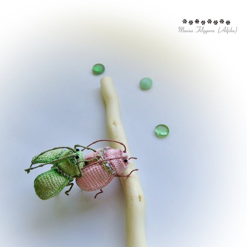 May include: Two crocheted bugs, one green and one pink, are perched on a white branch. The bugs have black eyes and are detailed with small crocheted accents. Three green glass stones are scattered around the branch.
