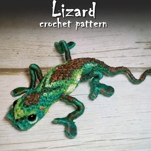 Op de afbeelding: Een groene, bruine en oranje gebreide hagedis met zwarte ogen. De hagedis ligt op een wit houten oppervlak. De tekst "Lizard crochet pattern" staat boven de hagedis.