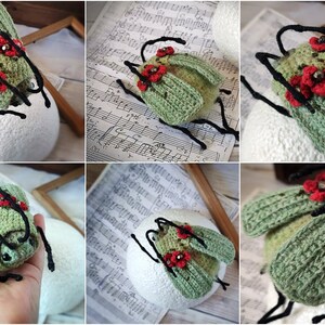 May include: A crocheted green beetle with red flower accents and black legs. The beetle is sitting on a white surface.