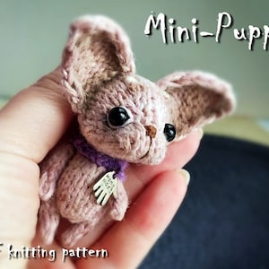 Puede incluir: Un pequeño cachorro de punto con ojos marrones y una bufanda morada. El cachorro sostiene una etiqueta plateada con las palabras "Hand Made" en ella. El cachorro está siendo sostenido en la mano de una persona. La imagen se titula "Mini-Puppy" y dice "PDF knitting pattern" en la parte inferior.