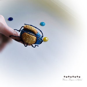 May include: A crocheted blue and yellow bug with black legs. The bug is sitting on a white surface.