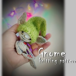 Könnte beinhalten: Handgestrickte Zwergenpuppe mit grünem Hut und grauem Körper. Die Puppe hat ein rotes Herz auf der Brust. Der Text "Gnome knitting pattern" ist in weißer Schrift geschrieben. Die Puppe wird in einer Hand gehalten.