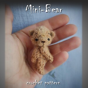 Puede incluir: Un pequeño oso de peluche de color marrón tejido a crochet con ojos negros, sentado en la palma de una mano. El texto "Mini-Bear crochet pattern" está debajo del oso.