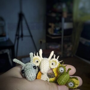 K&ouml;nnte beinhalten: Vier geh&auml;kelte Amigurumi-Spielzeuge mit gro&szlig;en Augen. Die Spielzeuge sind in verschiedenen Farben, darunter Gr&uuml;n, Wei&szlig; und Grau. Die Spielzeuge werden in der Hand einer Person gehalten.
