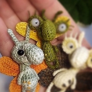 K&ouml;nnte beinhalten: Eine Sammlung kleiner geh&auml;kelter Tierfiguren. Die Spielzeuge sind ein gr&uuml;n-gelber Schmetterling, ein grau-oranger Schmetterling und ein wei&szlig;-cremefarbenes Wesen mit gro&szlig;en Augen.