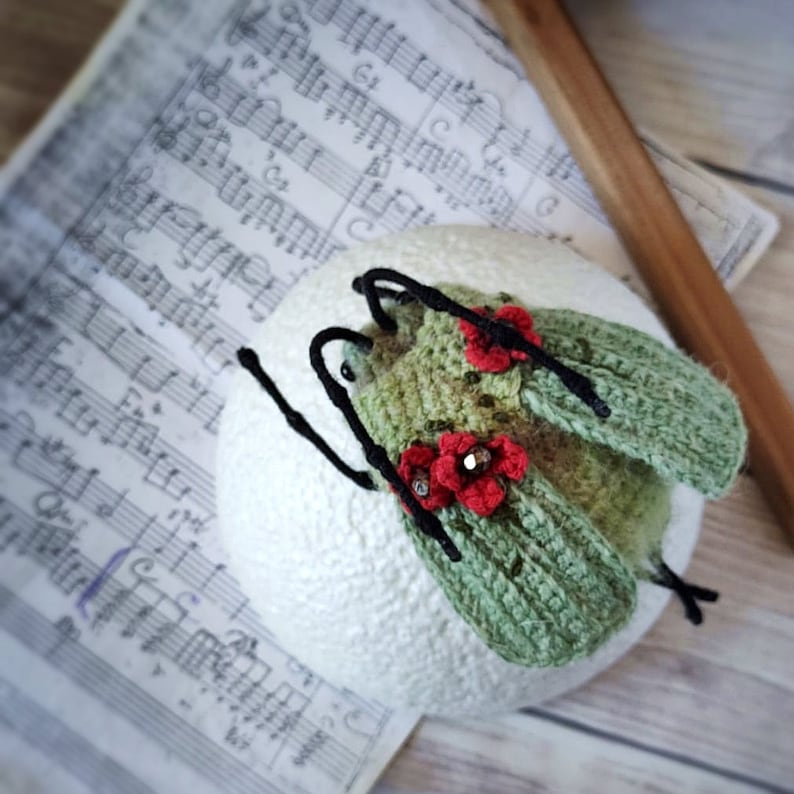 May include: A green crocheted insect with red flower accents and black wire legs. The insect is sitting on a white surface.