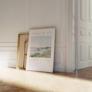 May include: A framed print of Psalm 23, featuring a landscape scene with a body of water and text. The print is leaning against a wall next to a canvas with a gold frame. The room has white walls and a wooden floor.