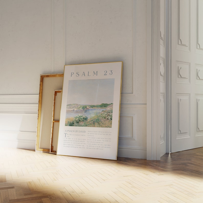 May include: A framed print of Psalm 23, featuring a landscape scene with a body of water and text. The print is leaning against a wall next to a canvas with a gold frame. The room has white walls and a wooden floor.