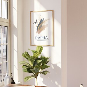 May include: A framed art print with a gold-colored frame hangs on a white wall. The artwork features abstract brushstrokes in navy blue, gold, and blush pink. The word "RAPHA" is printed in navy blue, with "THE GOD WHO HEALS" below it.