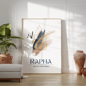 May include: A framed art print with abstract brushstrokes in navy blue, gold, and tan. The print features the word "RAPHA" and the phrase "THE GOD WHO HEALS" in a dark blue font. The artwork is displayed in a bright room with a plant and decorative vases.