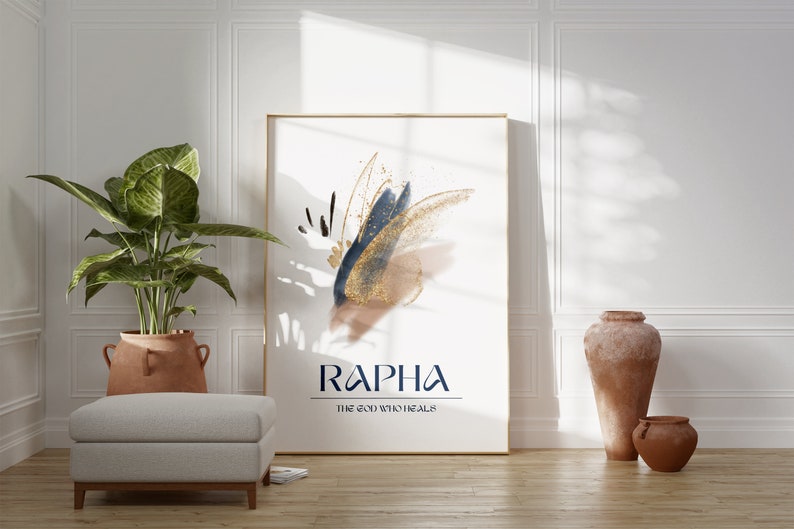 May include: A framed art print with abstract brushstrokes in navy blue, gold, and tan. The print features the word "RAPHA" and the phrase "THE GOD WHO HEALS" in a dark blue font. The artwork is displayed in a bright room with a plant and decorative vases.
