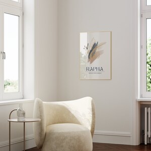 May include: A framed art print with abstract brushstrokes in gold, navy, and tan hangs on a white wall. The print is labeled "RAPHA" with the text "THE GOD WHO HEALS" below. A cream-colored armchair and small side table are in the foreground.