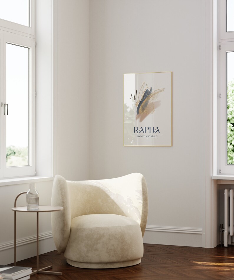 May include: A framed art print with abstract brushstrokes in gold, navy, and tan hangs on a white wall. The print is labeled "RAPHA" with the text "THE GOD WHO HEALS" below. A cream-colored armchair and small side table are in the foreground.