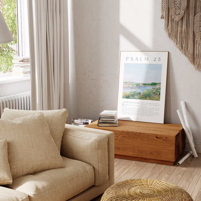 May include: A living room scene featuring a beige sofa with large cushions, a wooden coffee table, and a framed print with the text "Psalm 23." The print depicts a landscape with water and greenery. A woven ottoman and macrame wall hanging add texture.