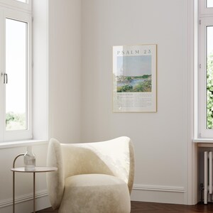 May include: A framed print of Psalm 23 hangs on a white wall. The print features a landscape scene with text at the top and bottom. A cream-colored armchair sits below the print. A small side table with a glass bottle and a stack of books is next to the chair.