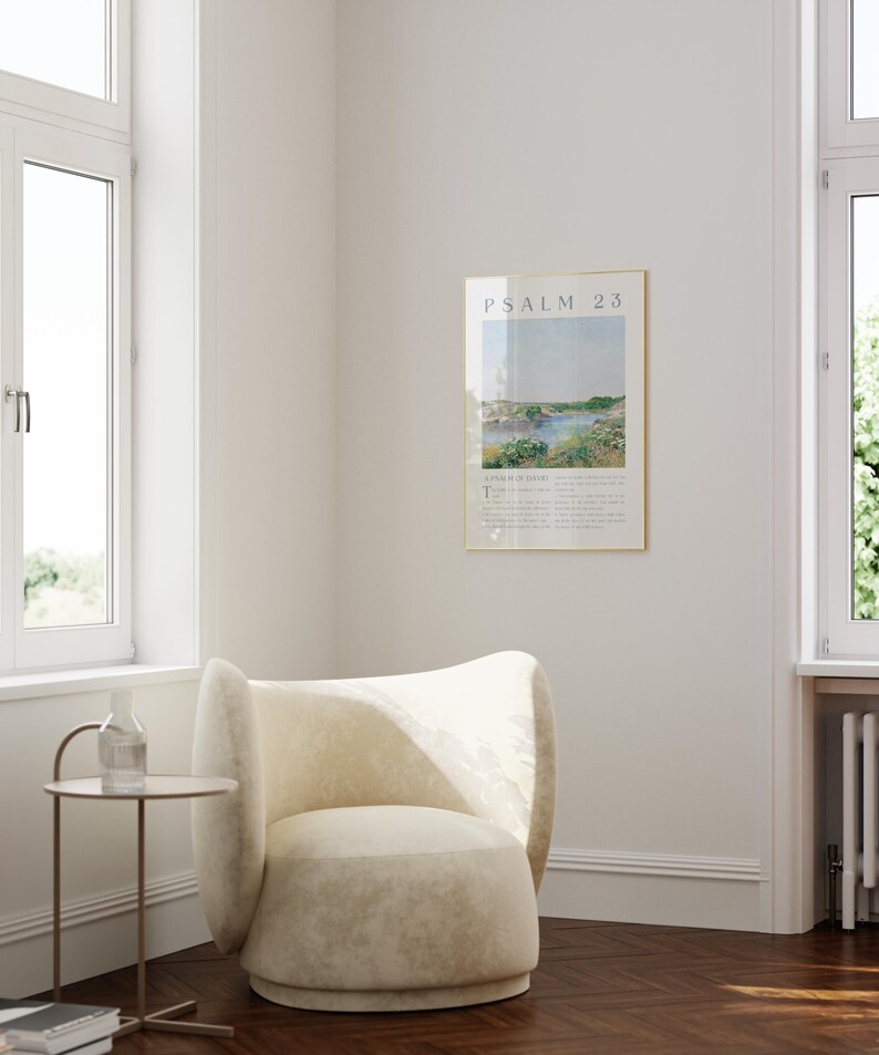 May include: A framed print of Psalm 23 hangs on a white wall. The print features a landscape scene with text at the top and bottom. A cream-colored armchair sits below the print. A small side table with a glass bottle and a stack of books is next to the chair.