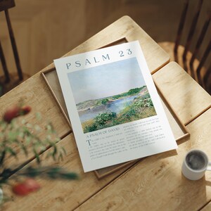May include: A framed print of Psalm 23, featuring a landscape painting of a river and green banks under a blue sky. The text "Psalm 23" is at the top, with "A Psalm of David" below the title. A small white mug sits on the table.