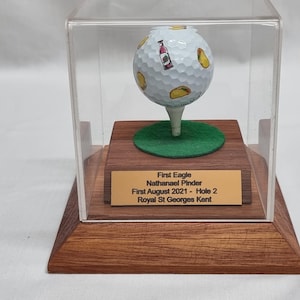 May include: A golf ball with food illustrations, displayed on a tee within a clear acrylic case. The ball rests on a green felt circle atop a wooden base with a gold plaque. The plaque reads "First Eagle, Nathanael Pinder, First August 2021 - Hole 2, Royal St Georges Kent."