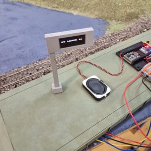 May include: A model train station sign with a digital display showing the text "OT LEAVE LL" mounted on a grey post. Next to the sign is a small black speaker connected by red and black wires to a circuit board and orange lever wire connectors.