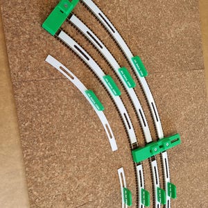 May include: Model train track pieces with white and black rails and green connectors. The pieces are arranged in curved and straight sections. The background is a corkboard surface.