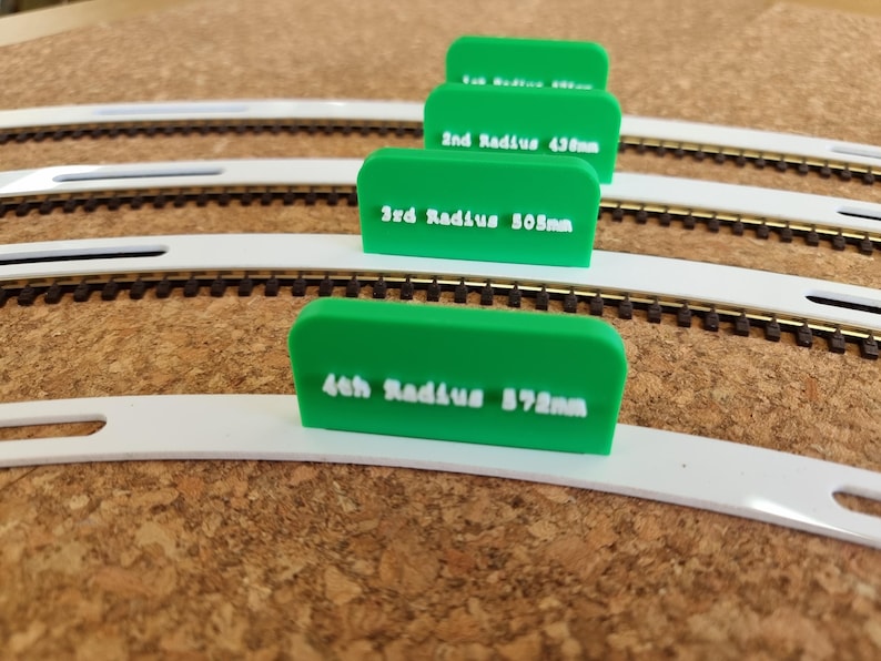 May include: Green plastic model train radius guides with white text indicating the radius in millimeters. The guides are placed on model train tracks. The text on the guides reads: 2nd Radius 438mm, 3rd Radius 508mm, and 4th Radius 572mm.