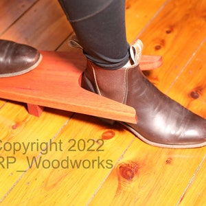 May include: A wooden boot jack with a brown leather boot on top. The boot jack is made of reddish-brown wood and has a cutout for the heel of the boot. The boot jack is sitting on a wooden floor. Copyright 2022 ERP_Woodworks