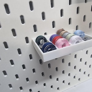 May include: A white pegboard with a white tray holding spools of thread in various colours. The thread colours include blue, purple, pink, green, and brown. The pegboard has evenly spaced oval holes.