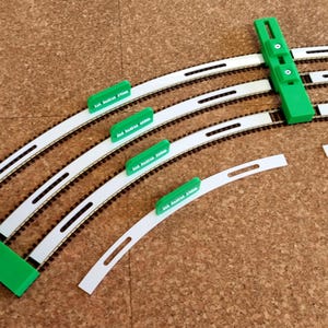 May include: A collection of model train track pieces, including curved track segments and green alignment tools. The tracks are white with black rails, and the alignment tools have text. The pieces are arranged on a brown surface.