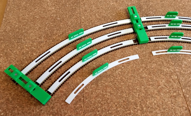 May include: A collection of model train track pieces, including curved track segments and green alignment tools. The tracks are white with black rails, and the alignment tools have text. The pieces are arranged on a brown surface.