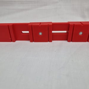 May include: A red plastic tool with two holes and a slot in the middle. The tool has two silver screws on the top.