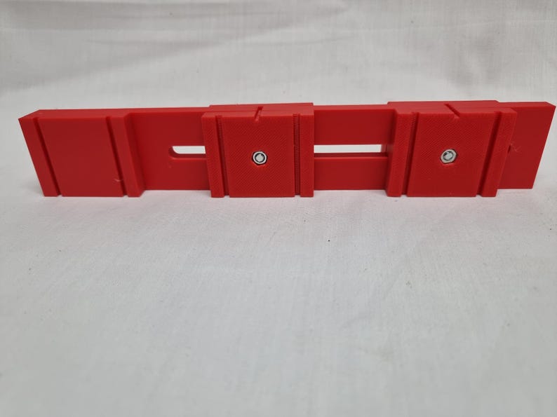 May include: A red plastic tool with two holes and a slot in the middle. The tool has two silver screws on the top.