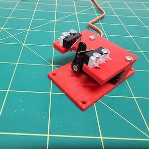 Pode incluir: Uma base vermelha impressa em 3D com dois microinterruptores pretos montados nela. A base tem um pequeno servo motor conectado a ela. O servo motor está conectado a um conjunto de fios.