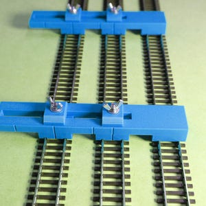 May include: Two blue plastic track gauges with adjustable knobs are placed on three sets of model train tracks. The tracks are on a green surface.