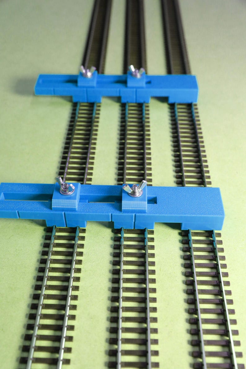 May include: Two blue plastic track gauges with adjustable knobs are placed on three sets of model train tracks. The tracks are on a green surface.