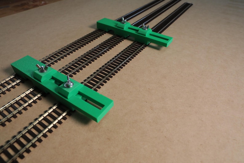 May include: Two green plastic track alignment tools with metal adjustment knobs. The tools are placed on top of three parallel train tracks.