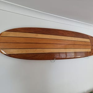 May include: A wooden surfboard with a striped design, featuring alternating light and dark brown wood panels. The surfboard is mounted on a white wall, showcasing its polished finish and craftsmanship. The board has a rounded nose and tail.